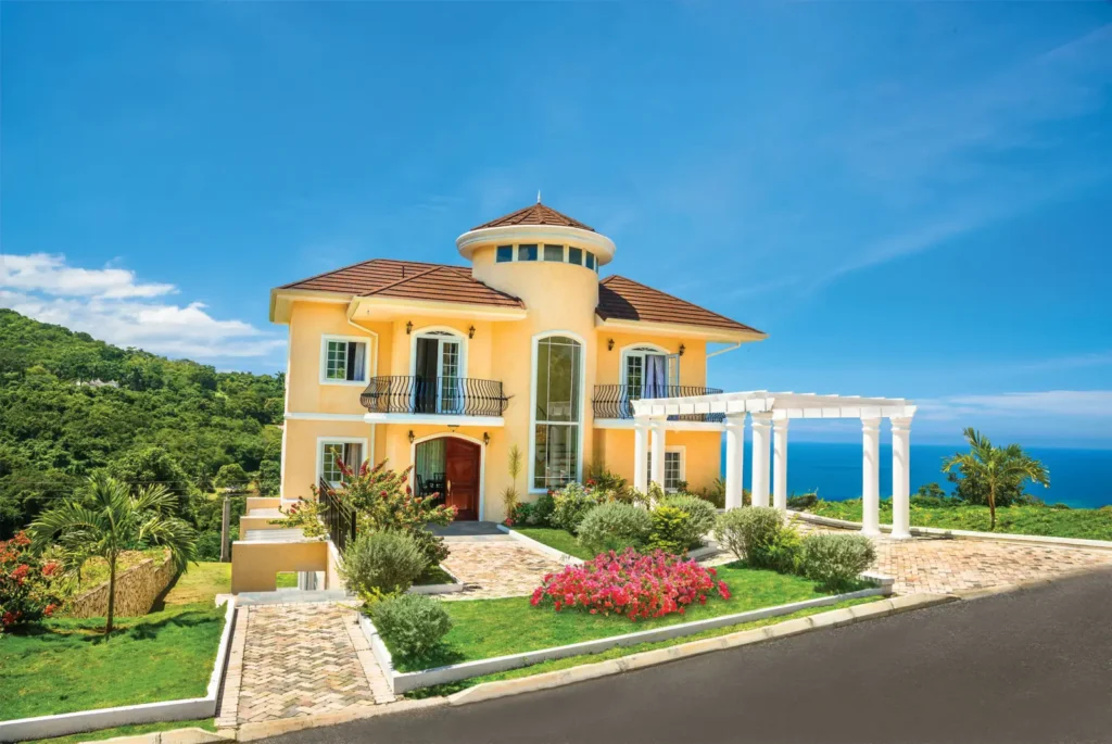 Yellow two-story villa with balconies, white pergola, landscaped garden and ocean view under a blue sky
