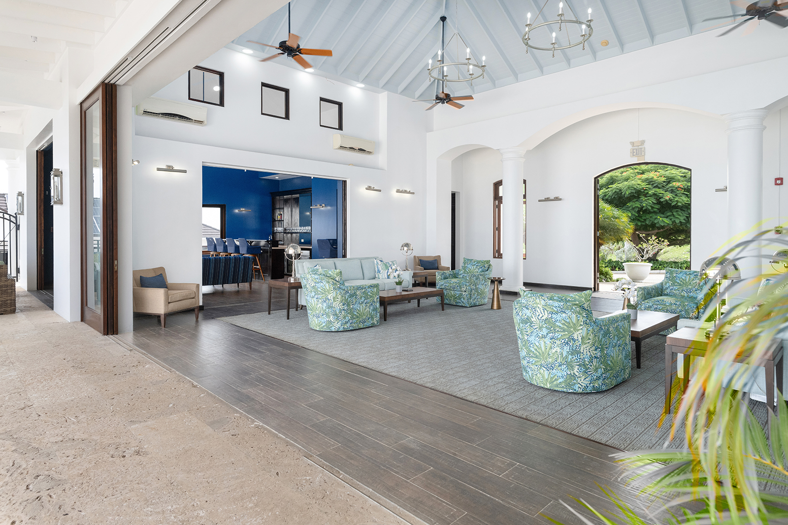 Bright clubhouse lounge with high vaulted ceiling, floral patterned armchairs, sofas, and open doorway to a garden