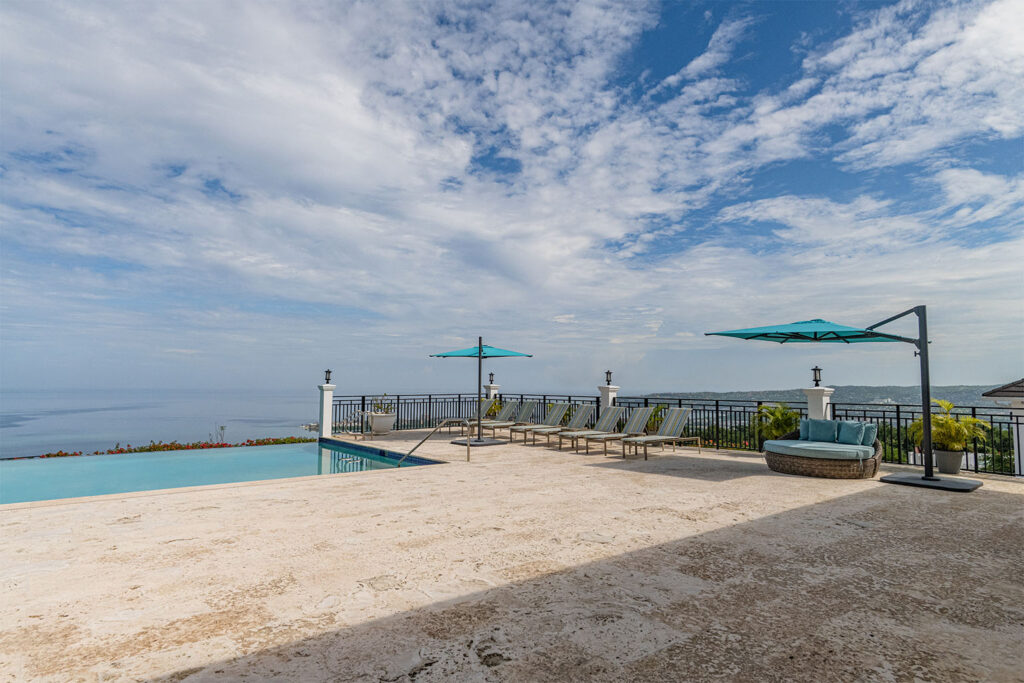 Infinity pool terrace with lounge chairs, turquoise umbrellas, and ocean view under a cloudy sky