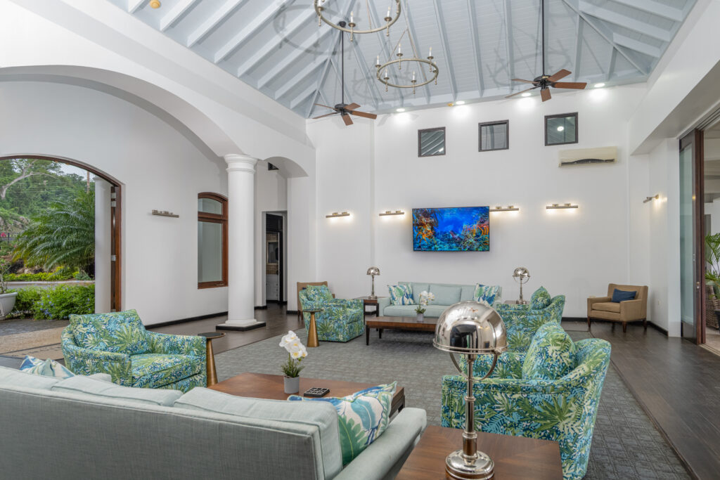Bright clubhouse lounge with vaulted blue ceiling, chandeliers, tropical-patterned seating, and wall-mounted TV